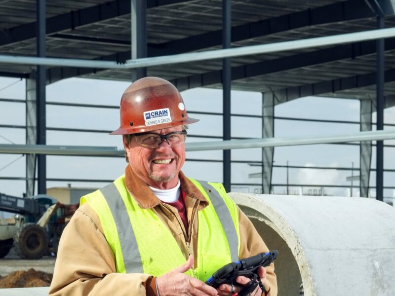 a Crain Construction employee smiling while holding an iPad on a construction site wearing a hard hat and safety vest