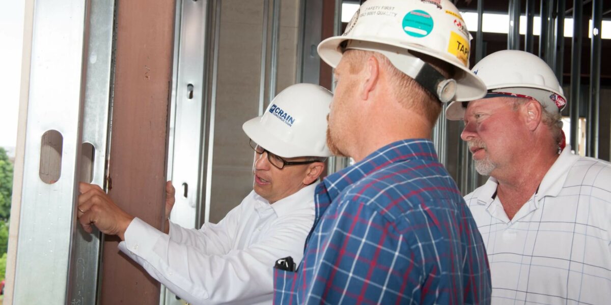 Three construction workers looking at the inside of a framed building with hard hats on.