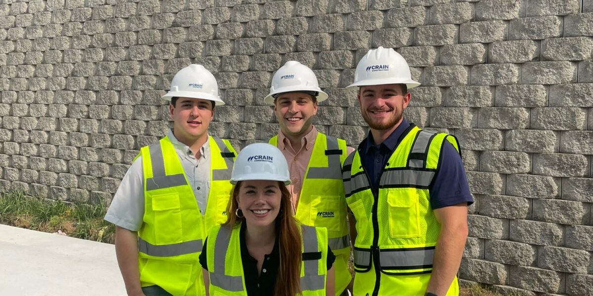 4 Crain Construction workers smiling in hard hats with neon vests on in front of a brick wall