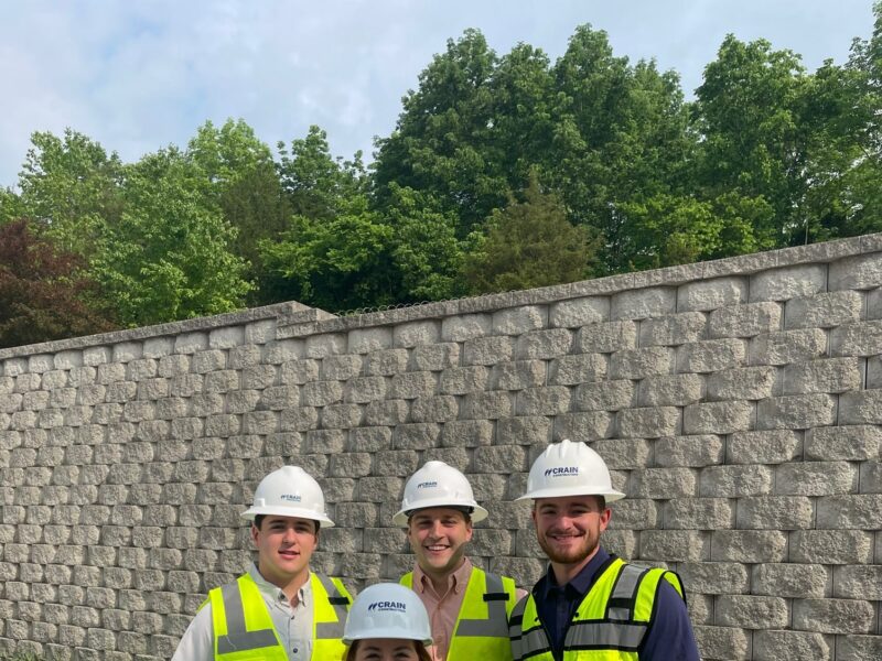 4 Crain Construction workers smiling in hard hats with neon vests on in front of a brick wall